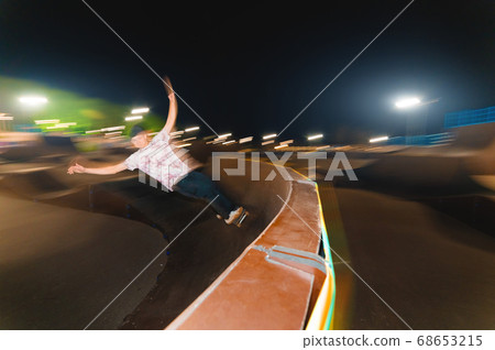 A young skateboarder rides at speed sideways along the ramp by spreading his hands to the sides. Night shot with long exposure at night speed in a skatepark 68653215