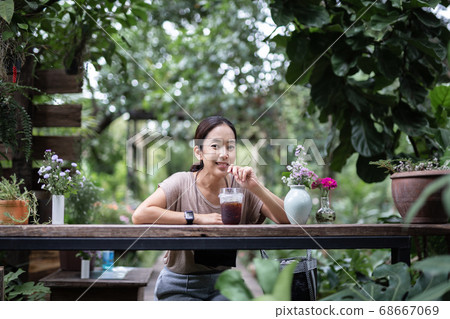 Woman resting outdoors sitting on wooden chair drinking iced coffee in nature garden. 68667069