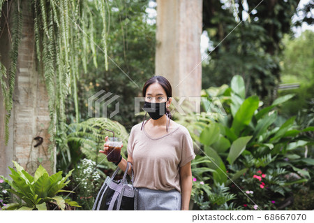 Woman in protective mask hand hold iced coffee walk in greenery park quarantine. 68667070
