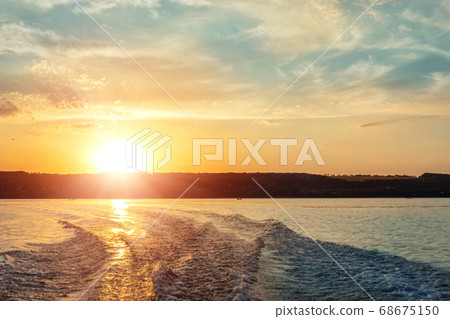Scenic bright vibrant blue to red warm sunset evening time landscape with motorbaot swirl trace on water surface of lake or river. Horizon coast line on background Scenic bright vibrant blue to red warm sunset evening time landscape with motorbaot swirl trace on water surface of lake or river. Horizon coast line on background 68675150