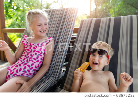 Two cute adorable caucasian blond little siblings enjoy having fun relaxing and eat fruit popsicle icecream sitting on sunbed in yard garden near pool sea beach. Brother and sister chilling outdoor Two cute adorable caucasian blond little siblings enjoy having fun relaxing and eat fruit popsicle icecream sitting on sunbed in yard garden near pool sea beach. Brother and sister chilling outdoor 68675252