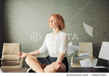 Serene business woman meditating in yoga lotus pose with flying papers around 68683984