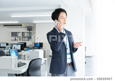 一個年輕的商人,通過在清晨獨自一人去辦公室與智能手機交談 一個年輕的商人,通過在清晨獨自一人去辦公室與智能手機交談 68689467