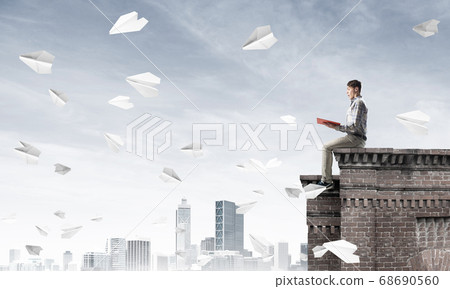 Handsome student guy on roof edge reading book and paper planes fly around Handsome student guy on roof edge reading book and paper planes fly around 68690560