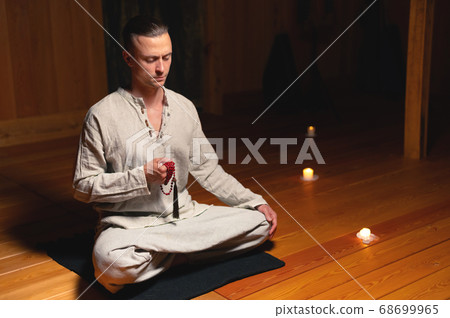 A portrait of an attractive Caucasian man sits in cotton robes in a lotus pose in a dark wooden practice room surrounded by candles A portrait of an attractive Caucasian man sits in cotton robes in a lotus pose in a dark wooden practice room surrounded by candles 68699965