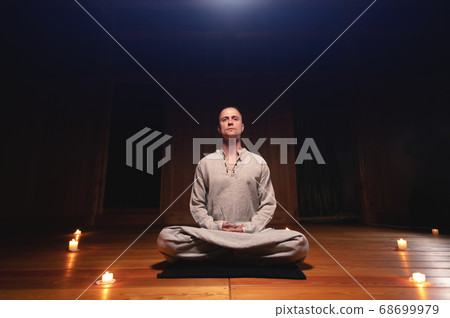 A portrait of an attractive Caucasian man sits in cotton robes in a lotus pose in a dark wooden practice room surrounded by candles 68699979