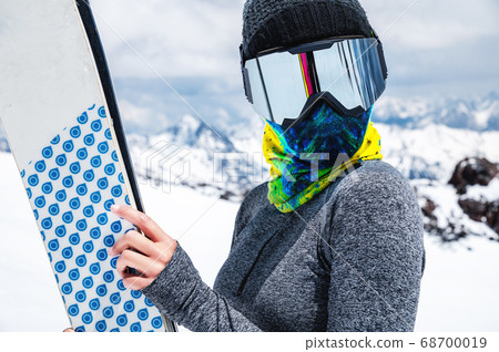 Portrait of a slender girl in a buff and balaclava in a ski mask and hat with a closed face next to skis on the background of snow-capped epic mountains. 68700019
