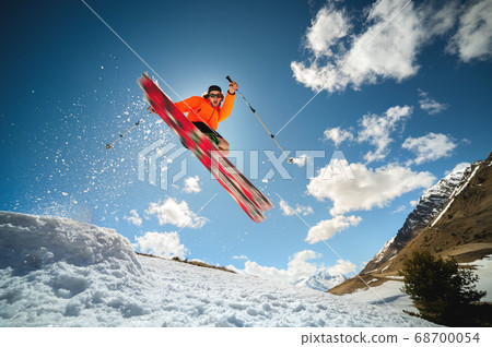A young stylish man in sunglasses and a cap performs a trick in jumping with a kicker of snow against the blue sky and the sun on a sunny day. The concept of park skiing in winter or spring 68700054