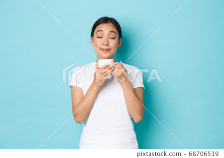 Portrait of delighted dreamy, pretty asian girl in white t-shirt, close eyes and smelling coffee, enjoying morning routine, sipping cappuccino from mug, standing light blue background Portrait of delighted dreamy, pretty asian girl in white t-shirt, close eyes and smelling coffee, enjoying morning routine, sipping cappuccino from mug, standing light blue background 68706519