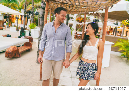 Summer vacation happy couple at tropical beach resort hotel walking to the beach. Young interracial people holding hands in casual clothes. Caribbean or Miami travel holiday lifestyle 68711937