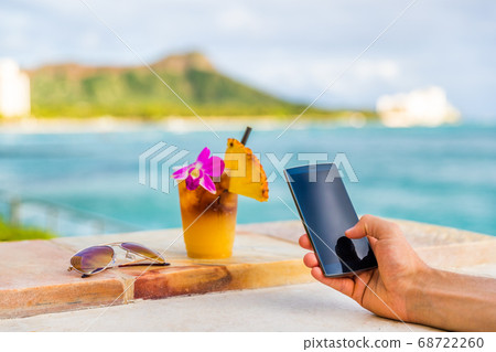 Phone app man texting taking mobile phone picture of Hawaii mai tai drink cocktail at Hawaii beach bar. Glass with flower, pineapple, sunglasses. Cellphone screen with diamond head, Honolulu vacation 68722260