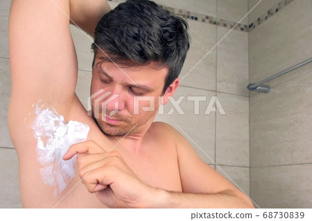 Portrait of man applying depilation cream on armpit using spatula in bathroom. 68730839