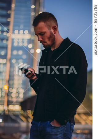 Dissatisfied man using smartphone texting. Male standing in the city in the evening outdoors using cell phone and waiting for taxi or uber. Young businessman looking at mobile phone Dissatisfied man using smartphone texting. Male standing in the city in the evening outdoors using cell phone and waiting for taxi or uber. Young businessman looking at mobile phone 68735768