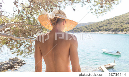 Rear view of topless beautiful woman wearing nothing but straw sun hat realaxing on wild coast of Adriatic sea on a beach in shade of pine tree. Rear view of topless beautiful woman wearing nothing but straw sun hat realaxing on wild coast of Adriatic sea on a beach in shade of pine tree. 68736344