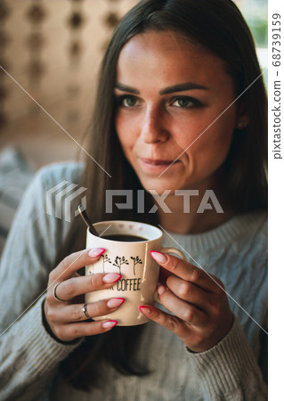 Close up portrait of a beautiful young woman drinking a cup of coffee by a window at home Close up portrait of a beautiful young woman drinking a cup of coffee by a window at home 68739159