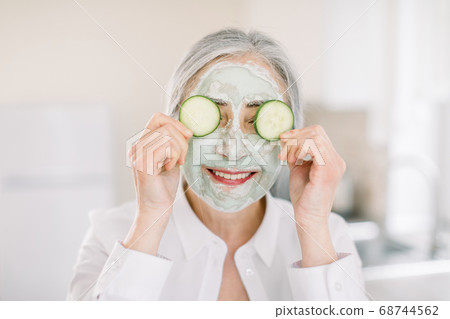 Close up of beautiful retired woman with facial mask on her face holding slices of fresh cucumber covering her eyes, on home interior background. Skin care and treatment, spa at home 68744562