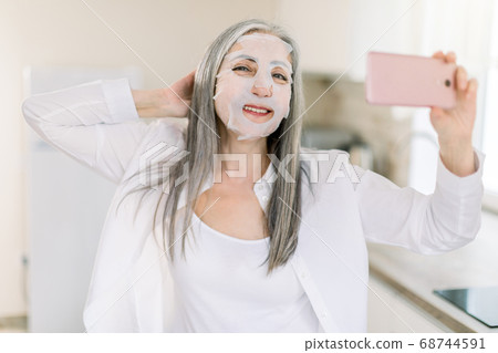 Beautiful retired grau haired lady in white shirt, standing at home kitchen and holding her cell phone, relaxing during beauty procedures with sheet cloth mask and smiling to camera 68744591