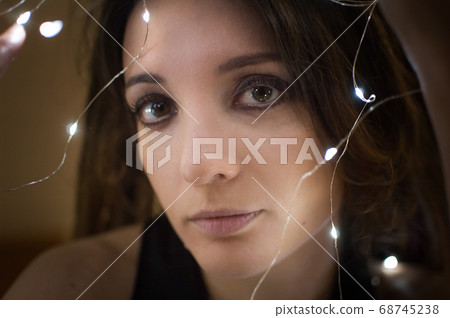 Indoors female portrait of a woman with dreadlocks hairstyle and smoky eyes makeup holding lights garland near her face, femininity and hollidays concepts Indoors female portrait of a woman with dreadlocks hairstyle and smoky eyes makeup holding lights garland near her face, femininity and hollidays concepts 68745238