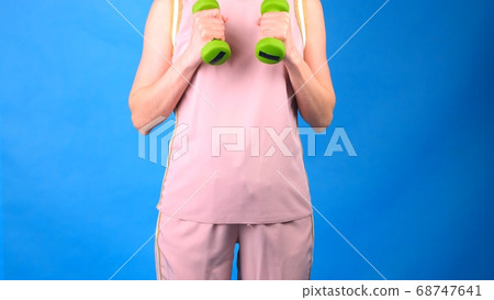 Female in a pink sports suit with dumbbells in hands does exercises on a blue background. The Female in a pink sports suit with dumbbells in hands does exercises on a blue background. The 68747641