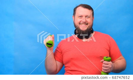 Funny fat man freaky in a wig and a pink t-shirt with dumbbells doing exercises on a blue background Funny fat man freaky in a wig and a pink t-shirt with dumbbells doing exercises on a blue background 68747652