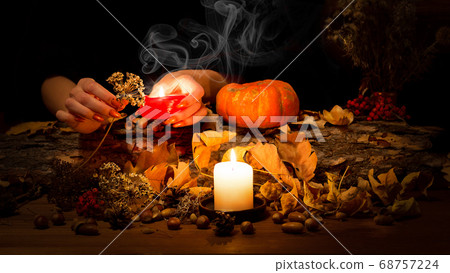 Altar of a pagan witch. Female hands with sharp red nails burn a dry herb in the candle fire. Pumpkin, nuts, dead leaves, acorns, ashberry background, selected focus, low key Altar of a pagan witch. Female hands with sharp red nails burn a dry herb in the candle fire. Pumpkin, nuts, dead leaves, acorns, ashberry background, selected focus, low key 68757224