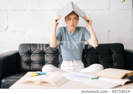 Unhappy redhead young woman sitting on couch and put the book on head. 68759014