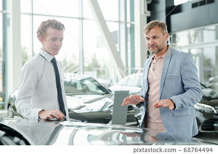 Mature male consumer in casualwear standing by one of new luxurious cars Mature male consumer in casualwear standing by one of new luxurious cars 68764494