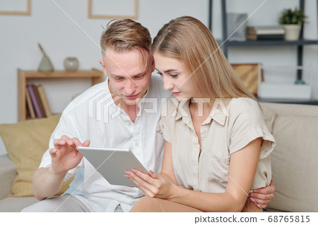 Contemporary young heterosexual couple using tablet while sitting on couch Contemporary young heterosexual couple using tablet while sitting on couch 68765815