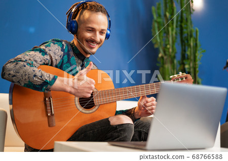 Young man sitting in his room and watching guitar tutorial on laptop Young man sitting in his room and watching guitar tutorial on laptop 68766588