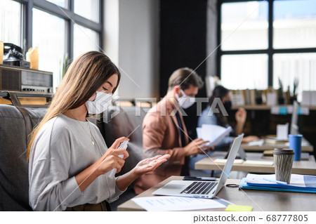 Portrait of young businesspeople with face masks working indoors in office, disinfecting hands. 68777805