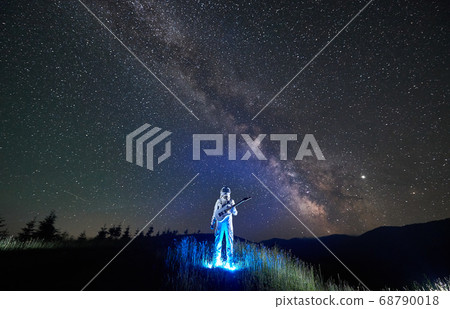 Illuminated spaceman fresh from the space standing in the middle of mountain meadow holding guitar, under Milky Way 68790018