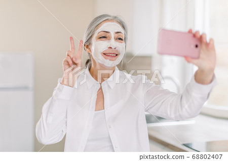 Portrait of positive, happy, smiling senior lady with long gray hair, with mud facial mask, having fun in kitchen at home, making photo on cell phone gesturing victory sign Portrait of positive, happy, smiling senior lady with long gray hair, with mud facial mask, having fun in kitchen at home, making photo on cell phone gesturing victory sign 68802407