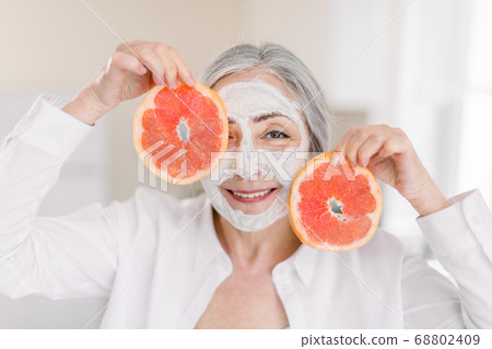 Joyful smiling senior woman in white shirt, with facial clay mask, posing to camera on home interior background with slices of fresh grapefruit. Beauty and Skin Care concept at Home Joyful smiling senior woman in white shirt, with facial clay mask, posing to camera on home interior background with slices of fresh grapefruit. Beauty and Skin Care concept at Home 68802409