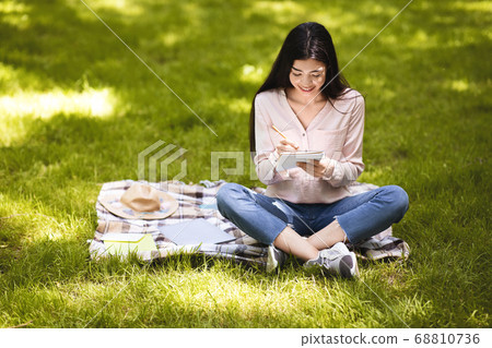Smiling asian girl making notes in notepad while sitting outdoors in park 68810736
