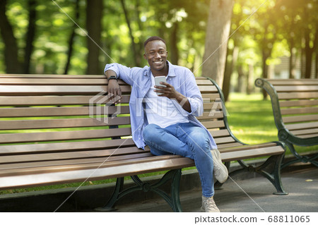 Smiling black man with mobile phone relaxing on bench at park on summer day 68811065