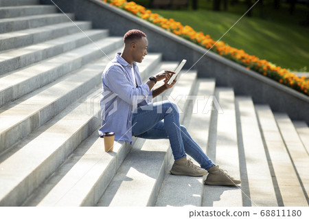 Side view of joyful black guy using tablet computer on stone stairs at city park Side view of joyful black guy using tablet computer on stone stairs at city park 68811180