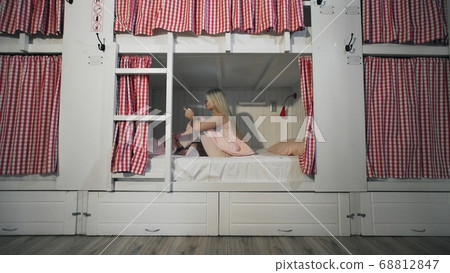 Young attractive woman sitting on the bed in the hostel room and using smartphone Young attractive woman sitting on the bed in the hostel room and using smartphone 68812847