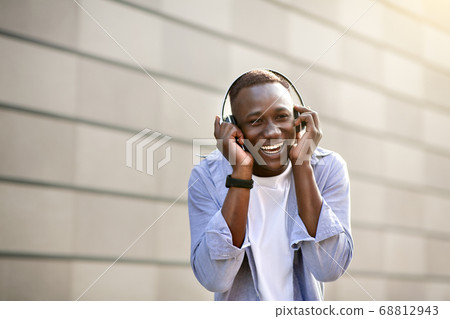 Laughing black guy listening to music in headphones near brick wall outside, blank space 68812943