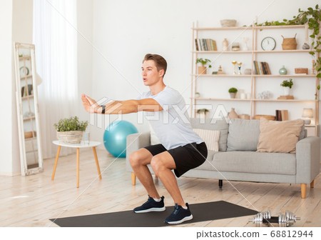 Keeps fit at home. Young man in sportswear and with fitness tracker squats in interior 68812944