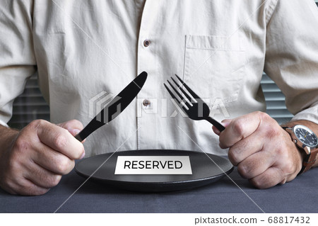 A man in a beige shirt at a table with a black empty plate. Black fork and knife. A man in a beige shirt at a table with a black empty plate. Black fork and knife. 68817432