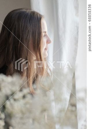 Portrait of young beautiful woman in black T-shirt by the window through white flowers. Sad, pensive girl is lost in her thoughts. Mindfulness concept. Portrait of young beautiful woman in black T-shirt by the window through white flowers. Sad, pensive girl is lost in her thoughts. Mindfulness concept. 68858336