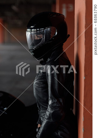 Stylish young woman motorcycle rider in black protective gear and full-face helmet near her bike on underground parking. 68863790