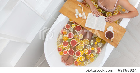Faceless female in bathtub with slices of oranges, lemon and grapefruits is reading the book. Woman having detox bath treatment. Pedicure tools on wooden tray. Faceless female in bathtub with slices of oranges, lemon and grapefruits is reading the book. Woman having detox bath treatment. Pedicure tools on wooden tray. 68866924