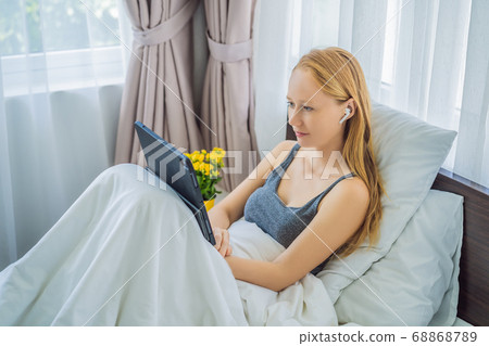 Young female lying in bed in morning, holding tablet computer, watching videos, reading news, looking relaxed and calm, enjoying free time 68868789