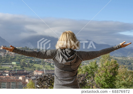 A blonde girl in a tracksuit looks from a hillside to a city in a valley and mountains with clouds in the distance A blonde girl in a tracksuit looks from a hillside to a city in a valley and mountains with clouds in the distance 68876972