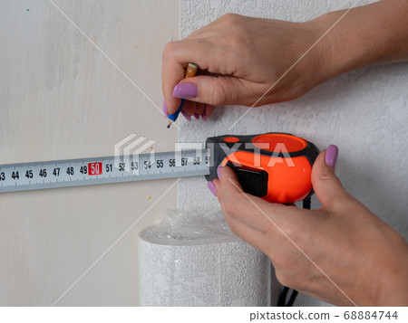 A woman makes measurements on the wall for wallpapering with a measuring ruler. A woman makes measurements on the wall for wallpapering with a measuring ruler. 68884744