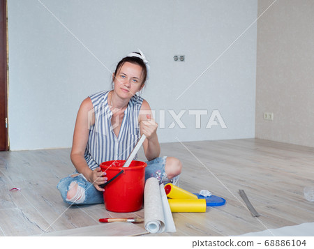 A woman stirs glue in a red bucket for wallpapering in an apartment. A woman stirs glue in a red bucket for wallpapering in an apartment. 68886104