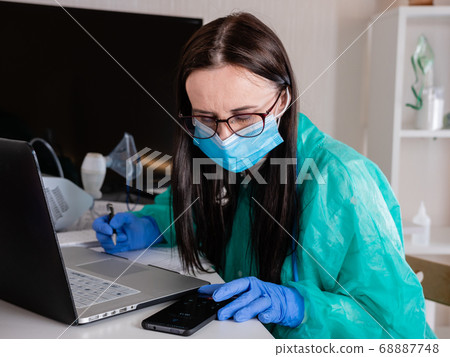A woman in a protective mask sits at the workplace and works with documents at the computer. A woman in a protective mask sits at the workplace and works with documents at the computer. 68887748