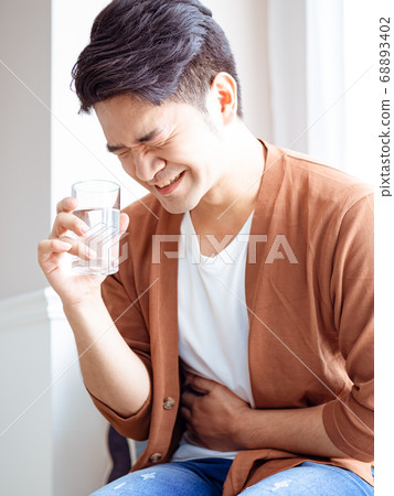 An adult sick man in a shirt with a glass of water and holds his hand on his stomach. An adult sick man in a shirt with a glass of water and holds his hand on his stomach. 68893402