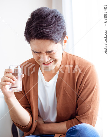 An adult sick man in a shirt with a glass of water and holds his hand on his stomach. An adult sick man in a shirt with a glass of water and holds his hand on his stomach. 68893403